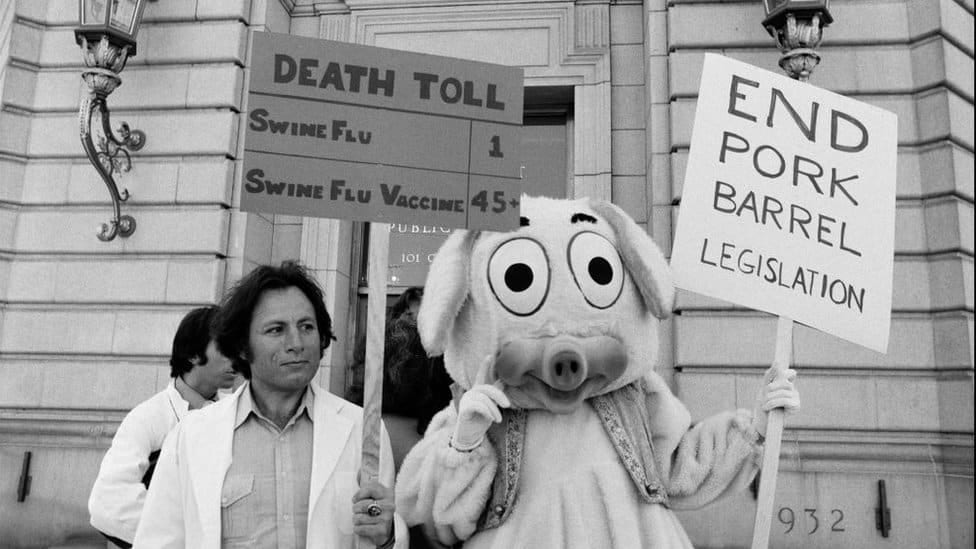 Protesters wearing white coats stand next to a protester in a pig costume, holding a sign saying: 
